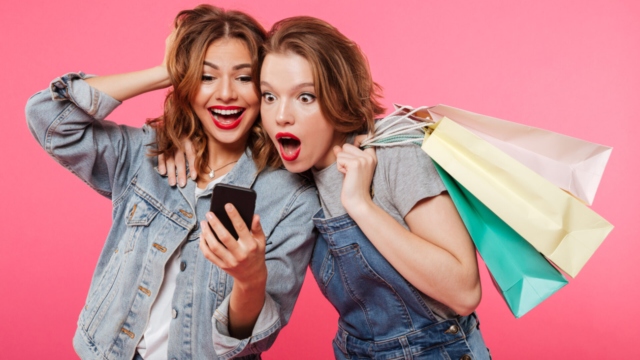 Image of shocked two women friends standing isolated over pink background. Looking aside holding shopping bags using mobile phone.