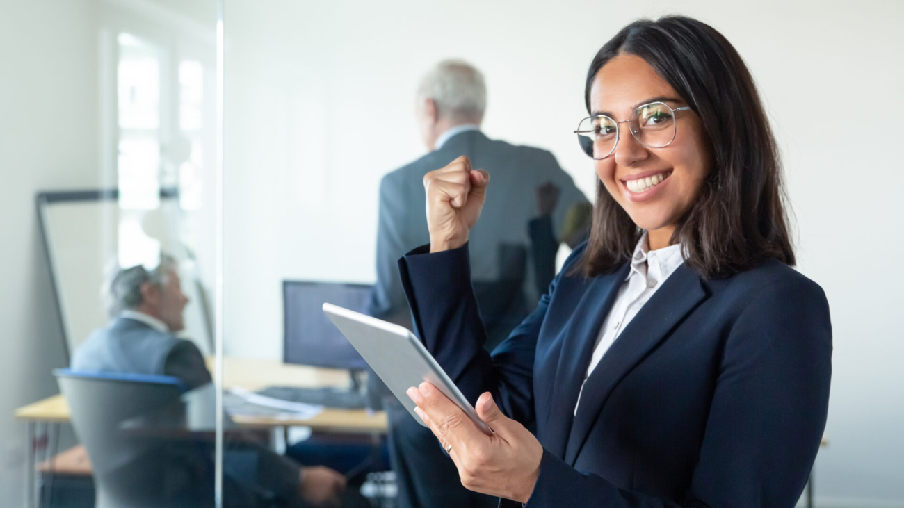 Happy female professional in glasses and suit holding tablet and making winner gesture while two businessmen working behind glass wall. Copy space. Communication concept
