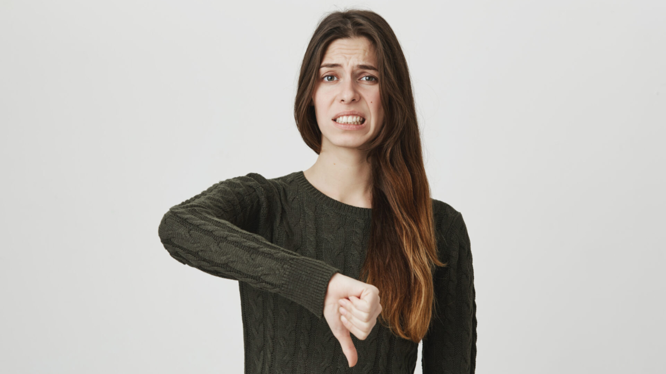 Portrait of dissatisfied frowning girl with long hair in casual clothes giving thumbs down gesture, expressing her disapproval and dislike of something. Negative emotions, reaction and feelings